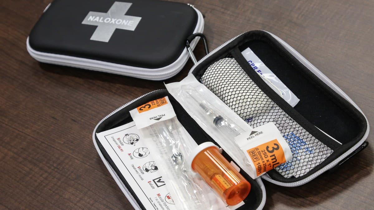 An open naloxone kit with a syringe, vials and instructions beside a closed black case marked Naloxone